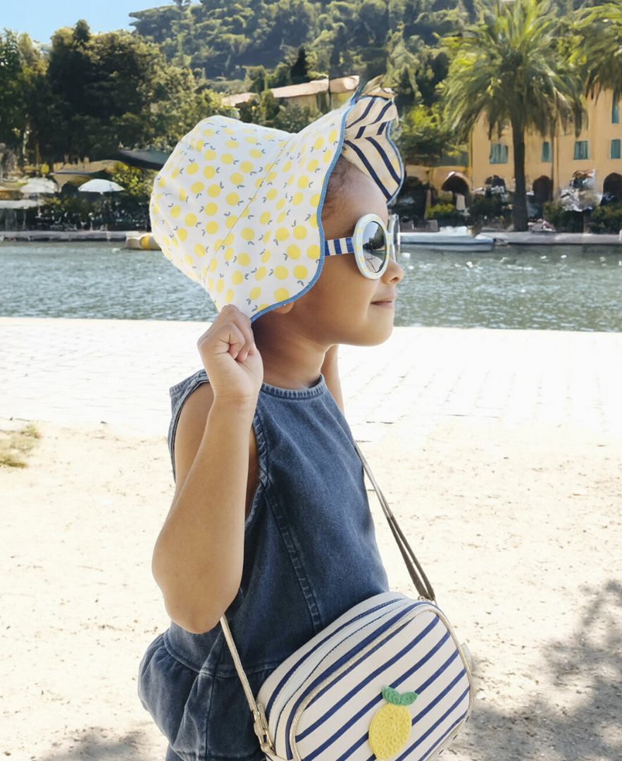ילדה מתוקה בכובע קייצי דו צדדי, משקפי שמש ותיק בעיצוב לימונים ופסים כחולים